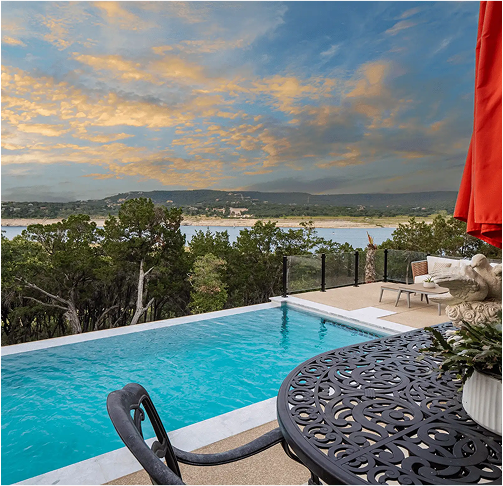 Infinity pool overlooking a lake with trees and a cloudy sky. Table and chairs on the patio.