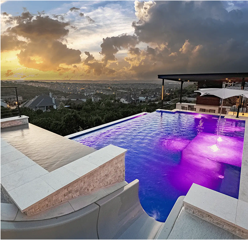 Rooftop pool with purple and blue lights overlooking a city at sunset with a cloudy sky.