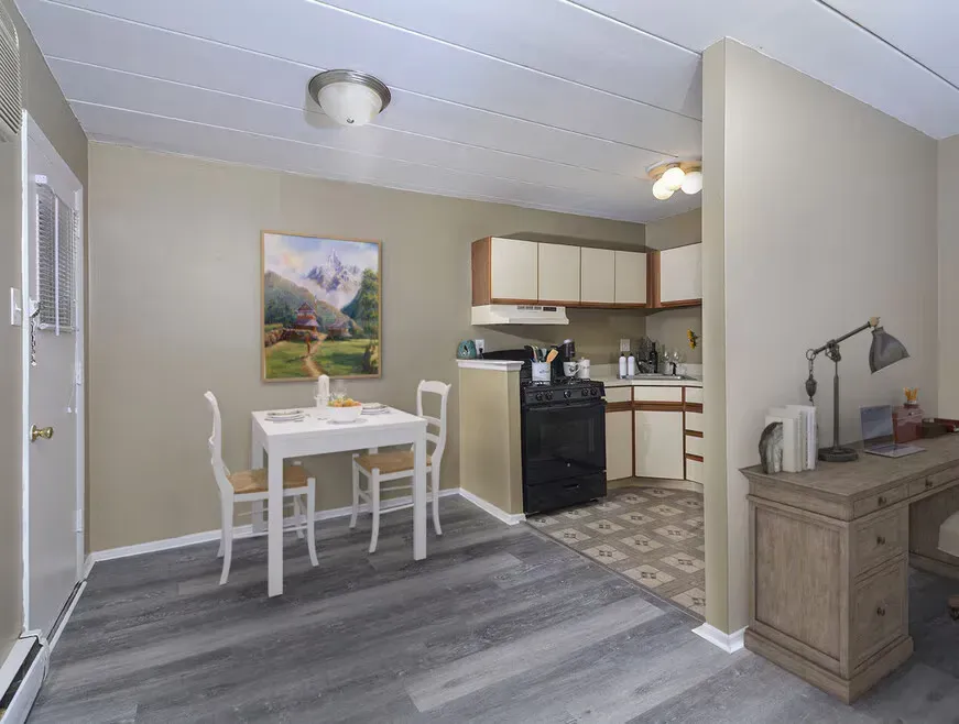 Small dining area beside a compact kitchen with white table, chairs, and gray carpeted floor
