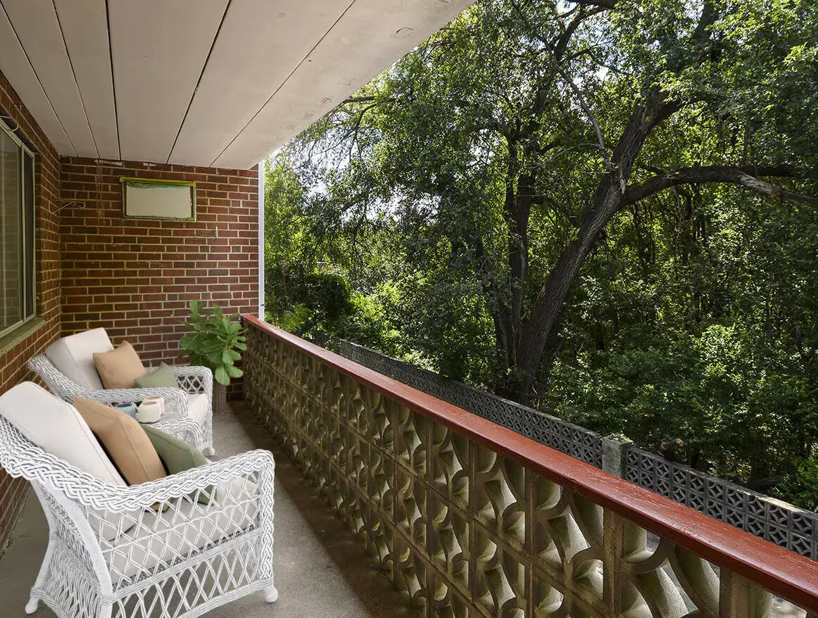 Covered balcony with white wicker chairs, brick wall, and lush green trees beyond the railing.