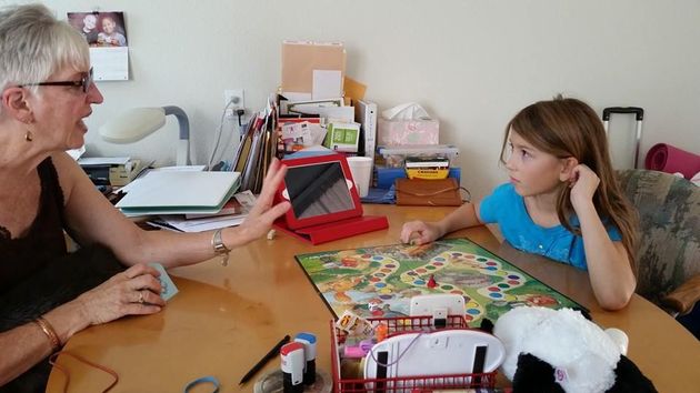 Grandmother and girl playing board game at a table, looking at each other. Red laptop, colorful game board.