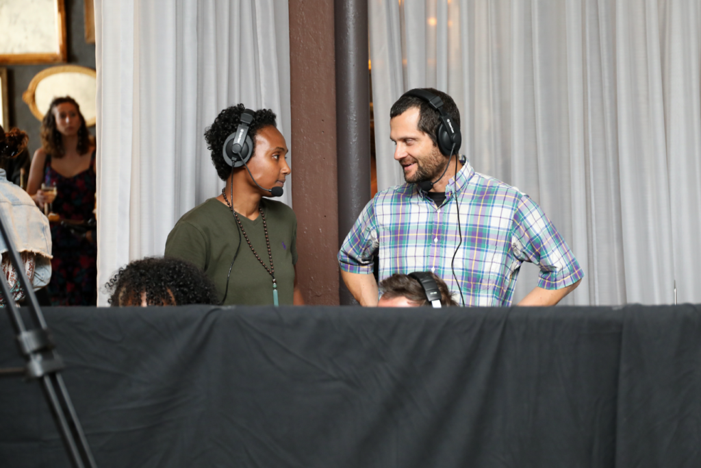 Two people with headphones behind a black barrier. A woman and a man appear to be talking.