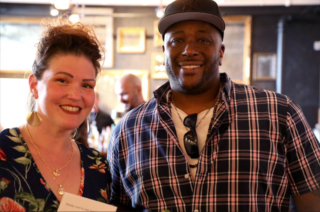 Woman and man smiling at the camera indoors. The man wears a cap and plaid shirt. The woman holds a paper.