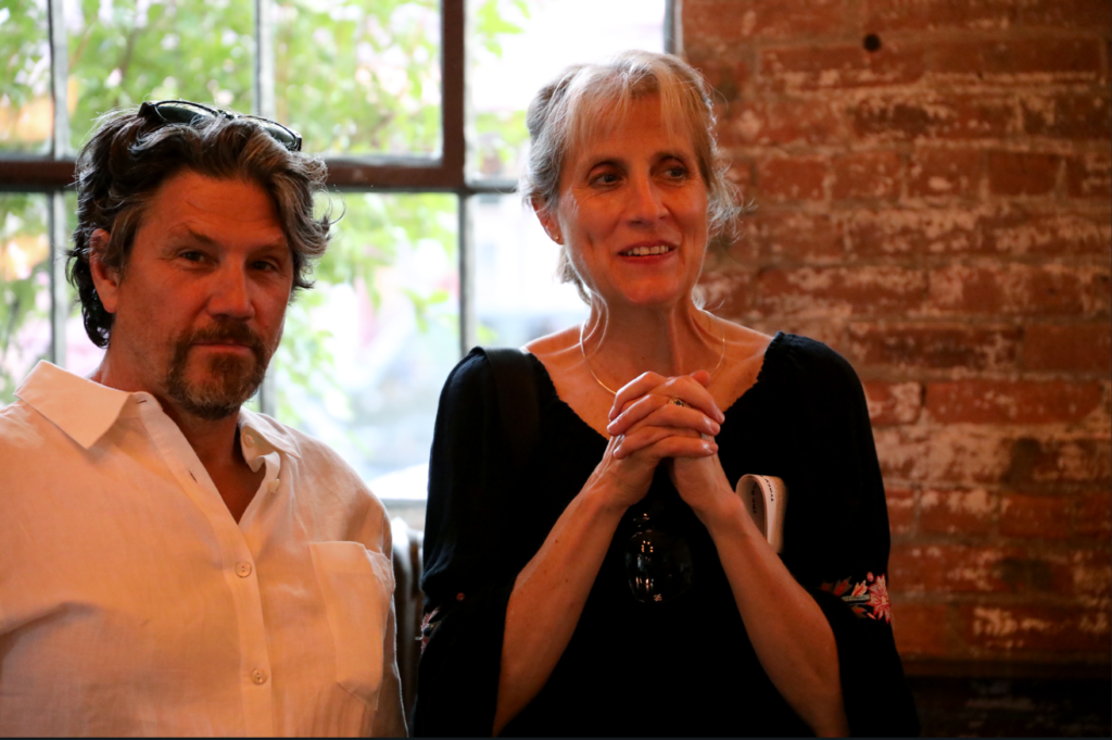 Man and woman sitting near a window and brick wall. The woman clasps hands, smiling. Man looks toward the camera.
