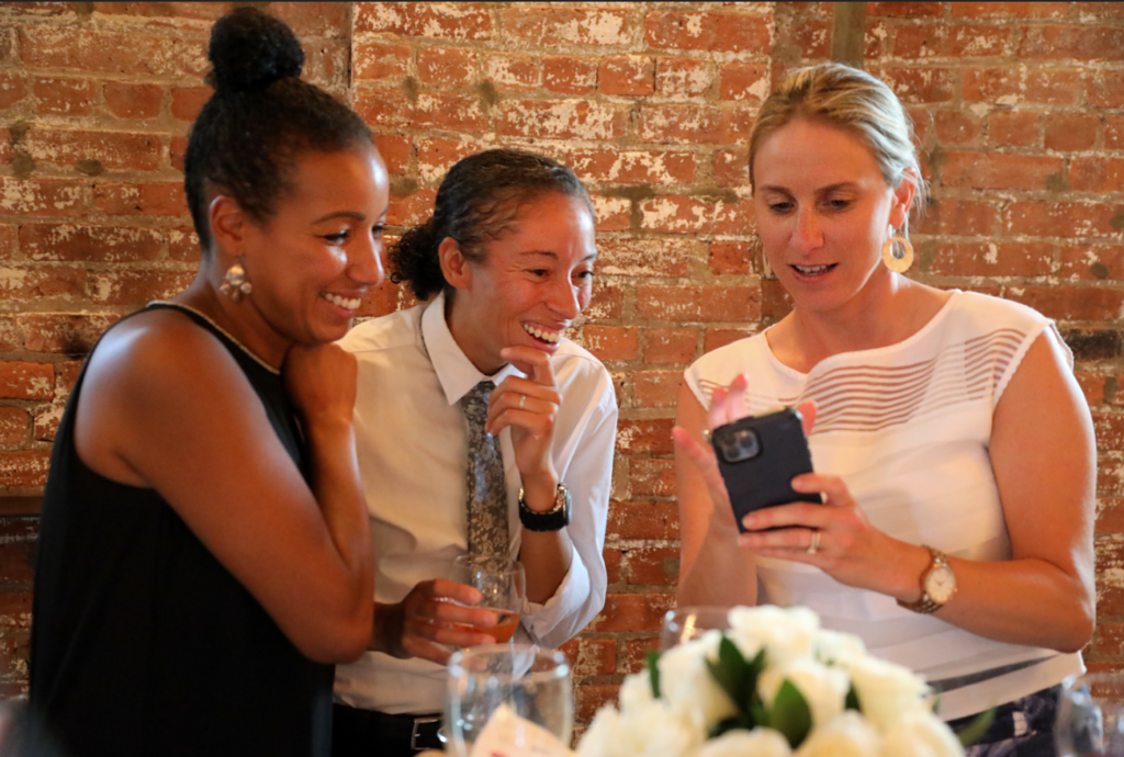 Three women smiling, looking at a phone. Brick wall in the background. One woman has a bun.