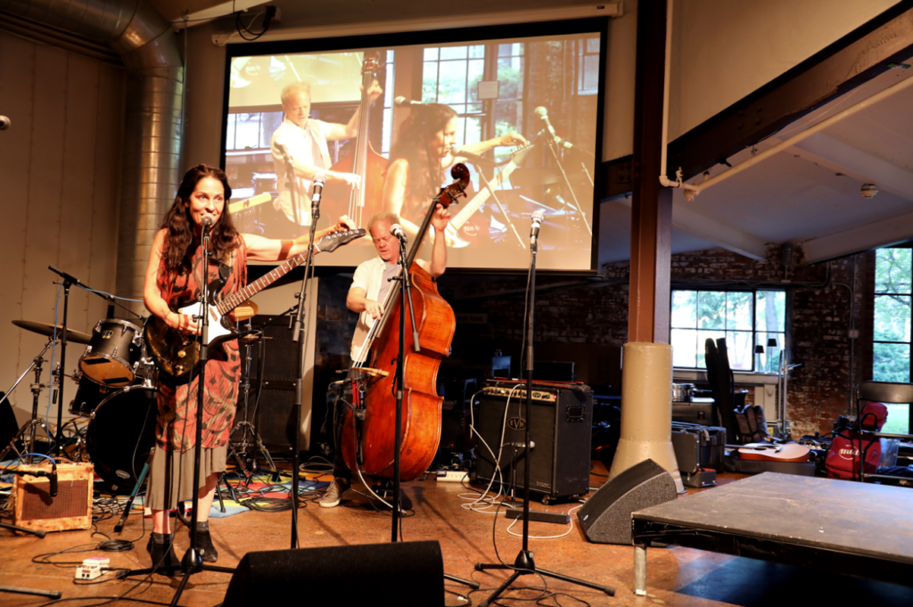 A band performs on stage: a woman guitarist, a bassist, with a screen showing musicians in the background.