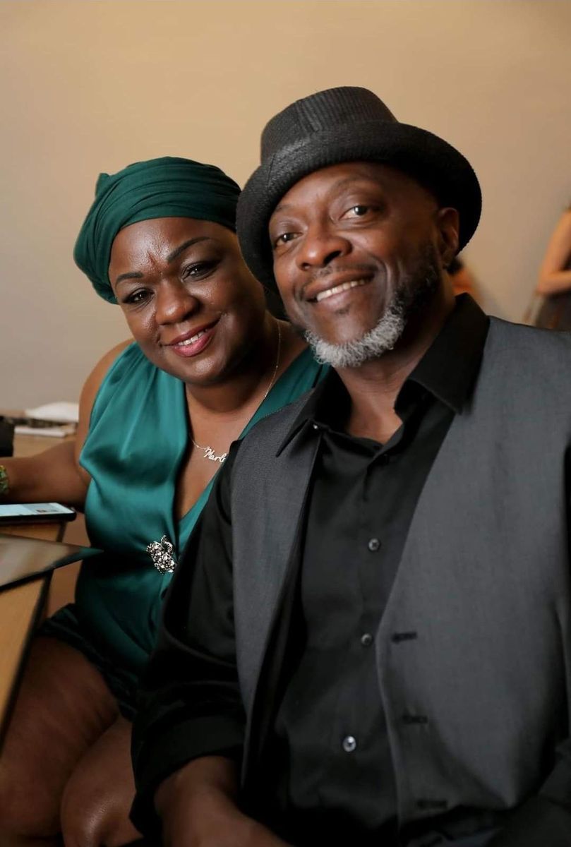Couple, woman in green headwrap and dress, man in fedora and vest, smiling, close up.