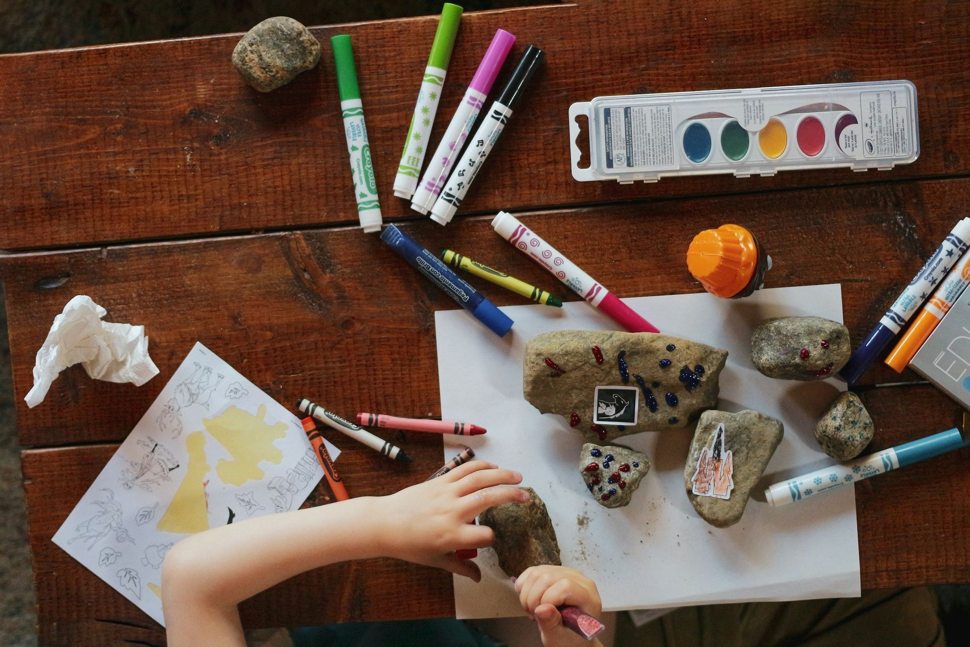 Child drawing on rocks with markers and crayons on a wooden table.