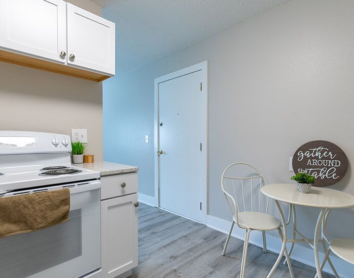 Small kitchen with white cabinets, stove, round table, and chair against light gray walls and wood-look floor