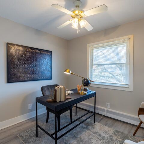 Small home office with black desk, lamp, wall chart, window, and ceiling fan on a light rug.