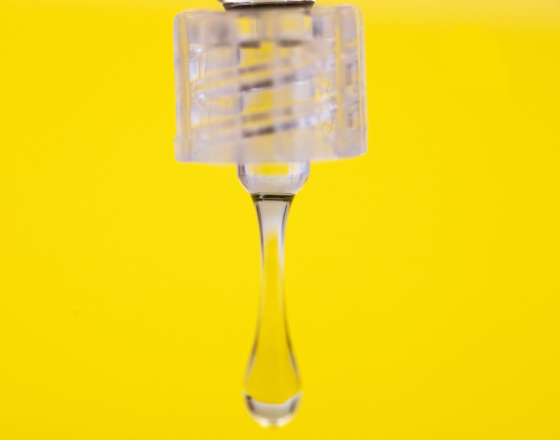 Clear liquid droplet hanging from a clear plastic device against a yellow background.