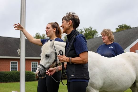 3 Riding Instructors standing next to a white horse smiling and laughing