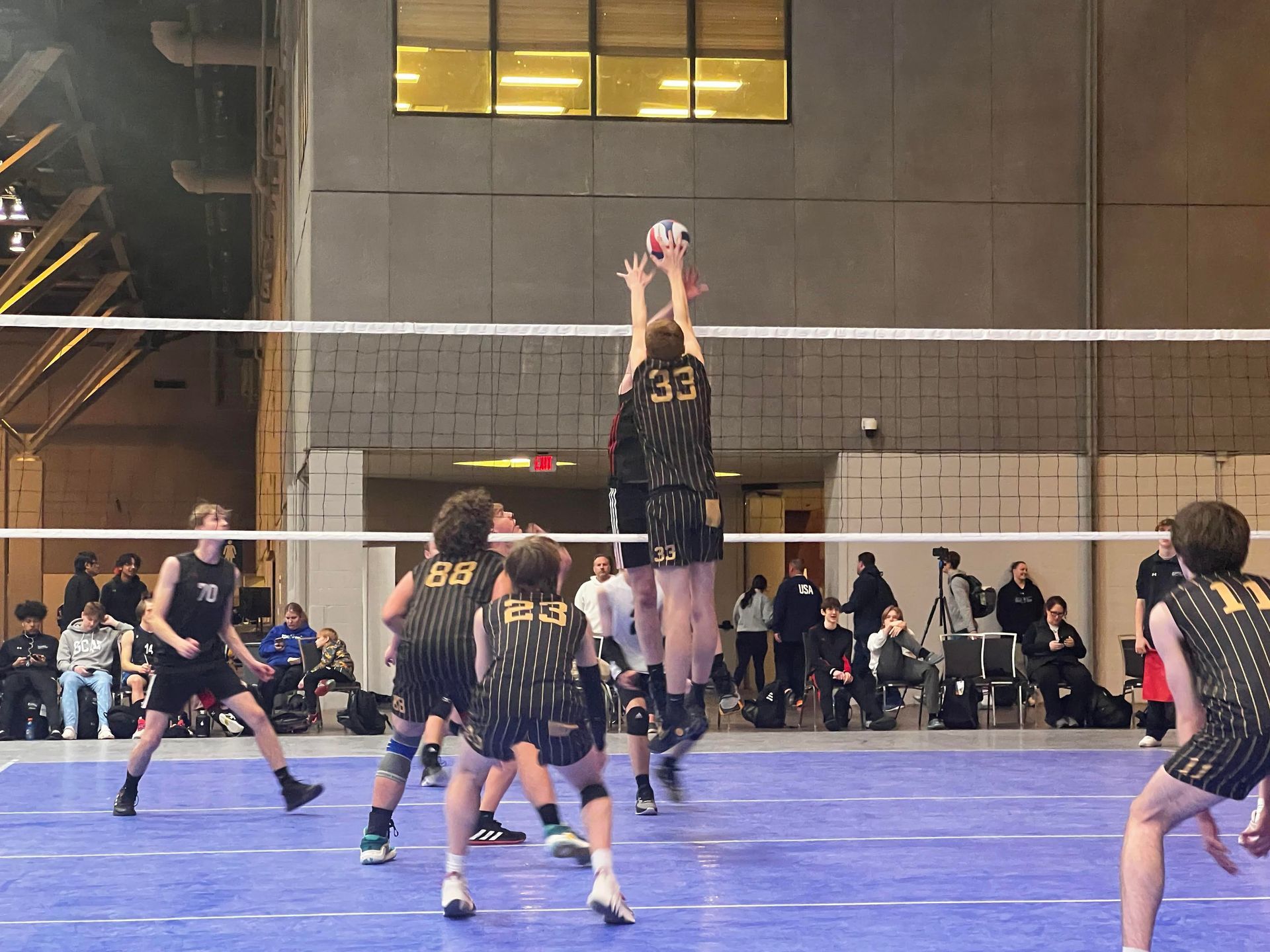 Boys' volleyball game; player in black jersey jumps to block ball at net. Indoor court, spectators.