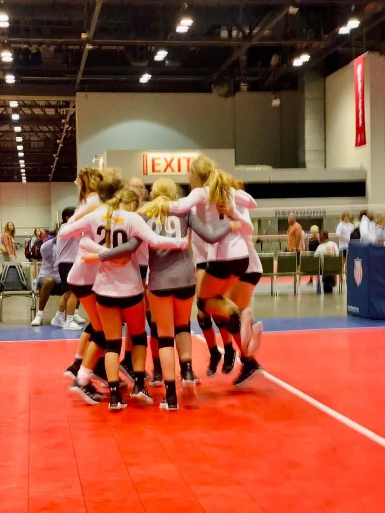 Volleyball team huddled on a red court, celebrating. They are wearing pink shirts, black shorts, and knee pads.