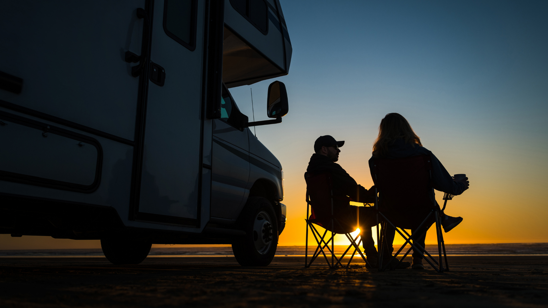 Two people in camping chairs watching sunset beside an RV on a beach
