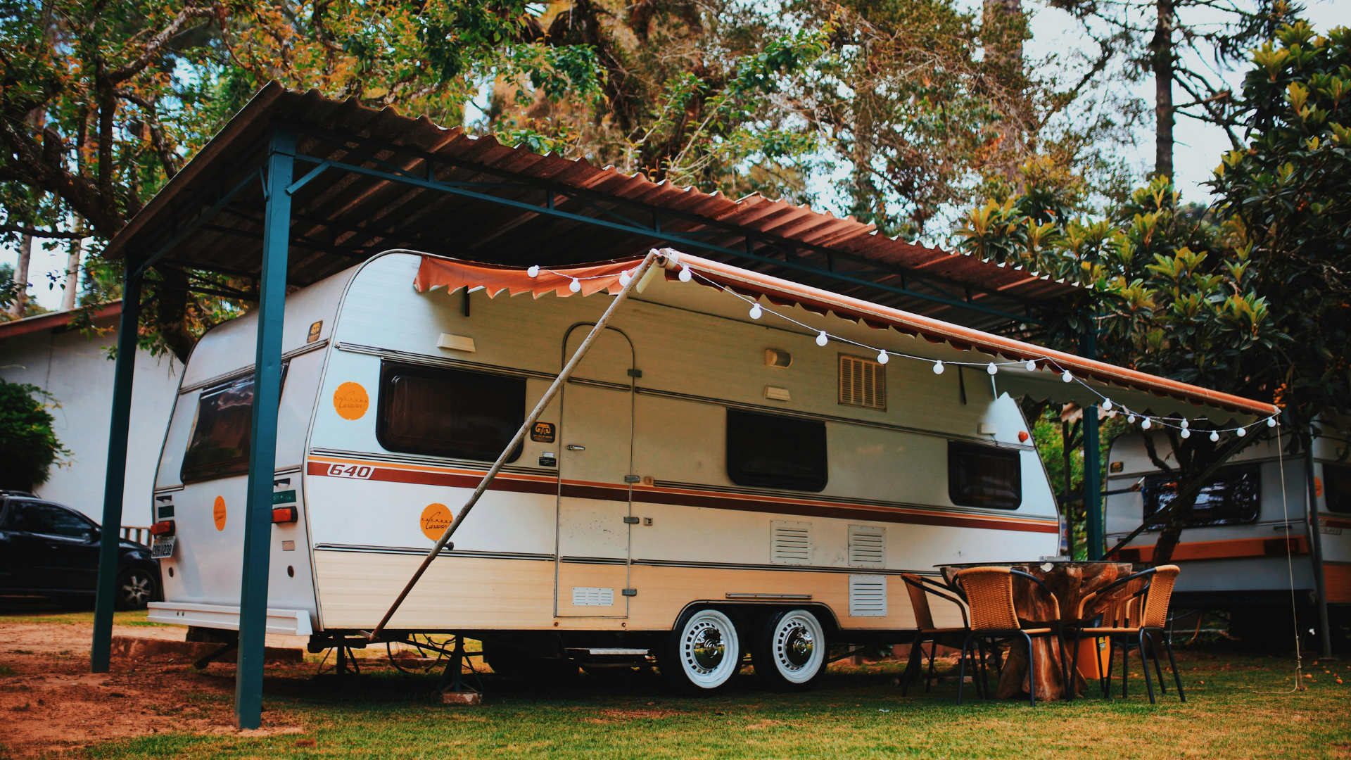 White camper trailer under a red-roof carport in a wooded campsite.