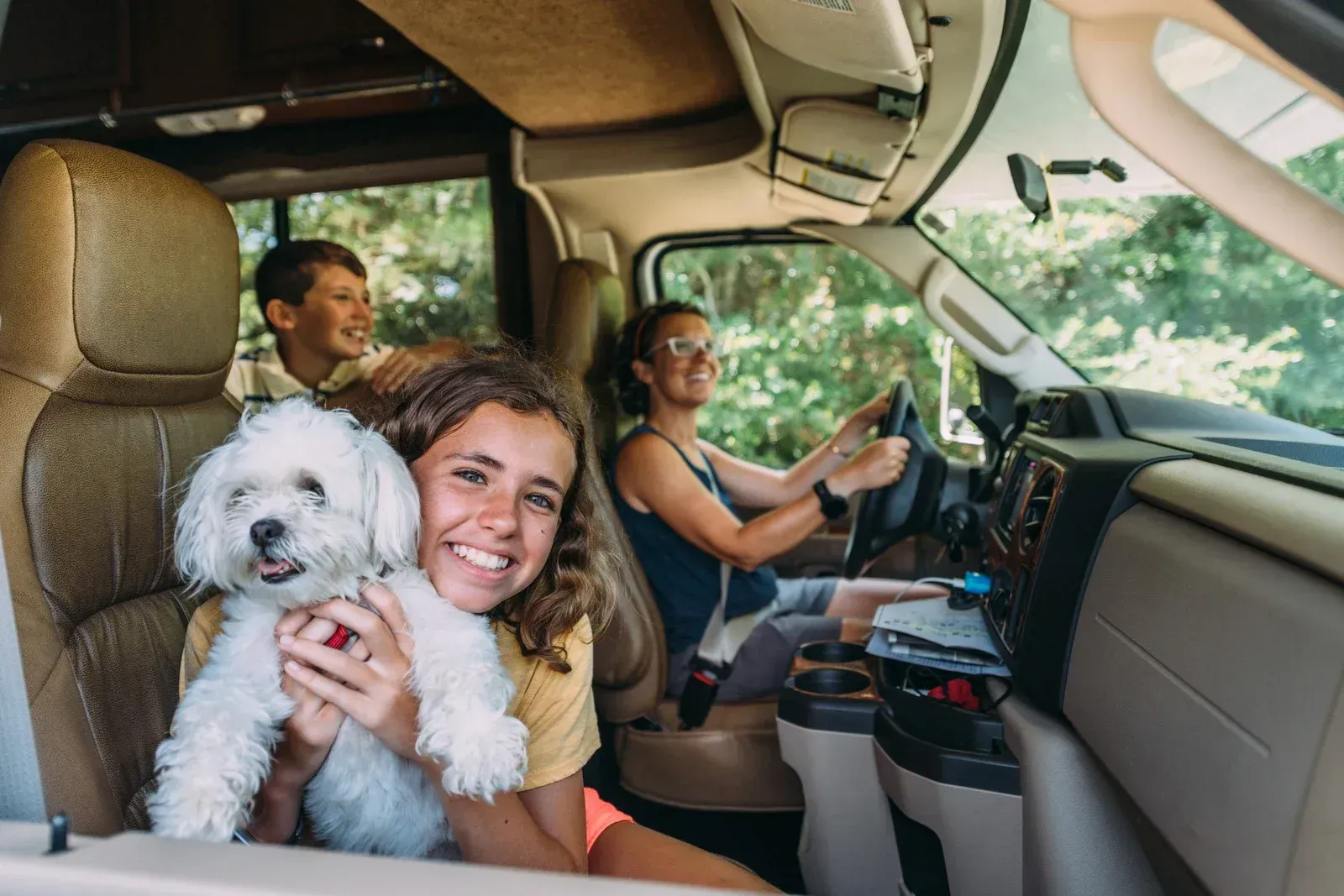 Three people and a white dog smiling inside a camper van, with one person driving.