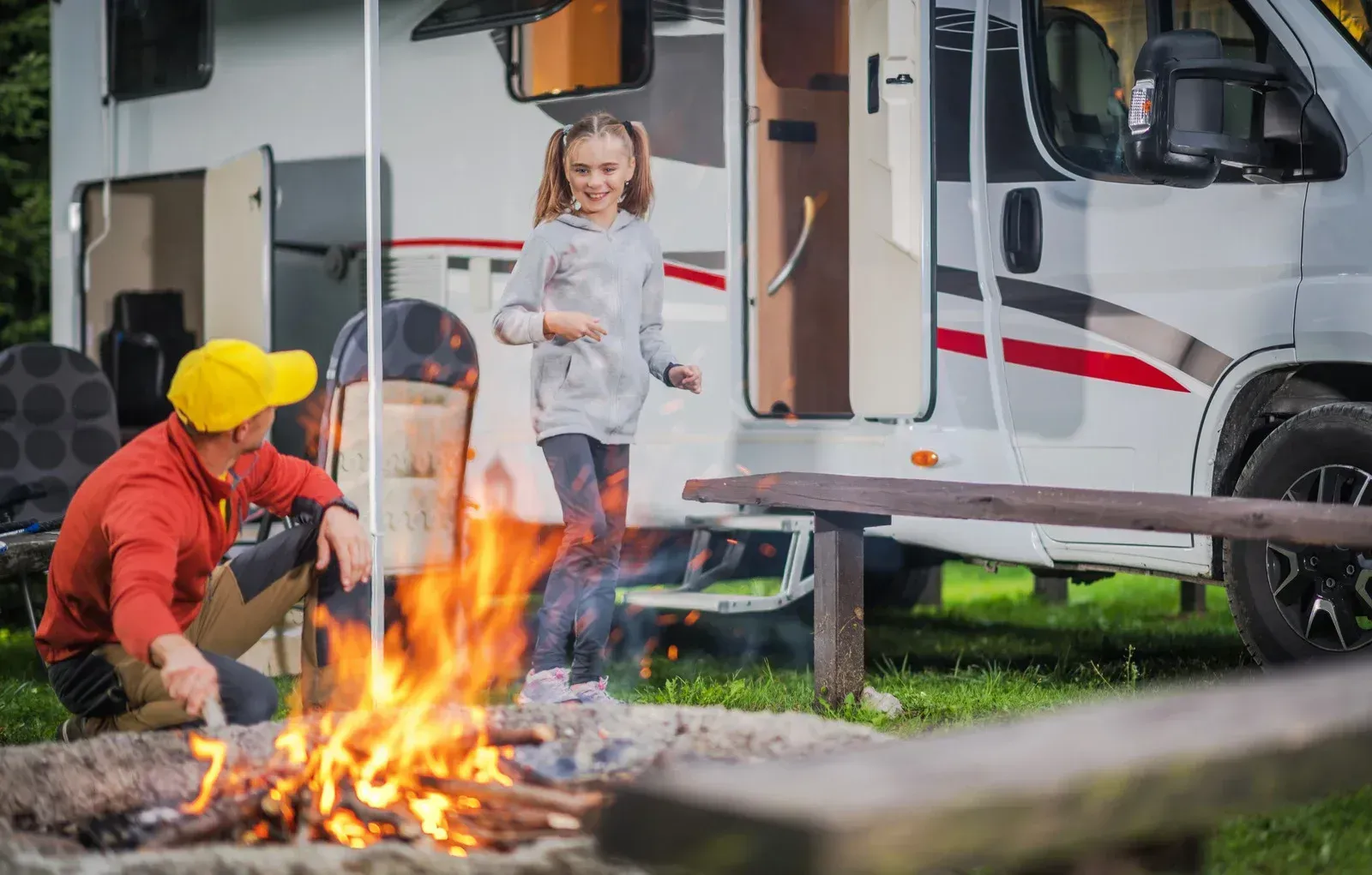 Child near a campfire outside a white RV, with an adult seated nearby and tents in the background