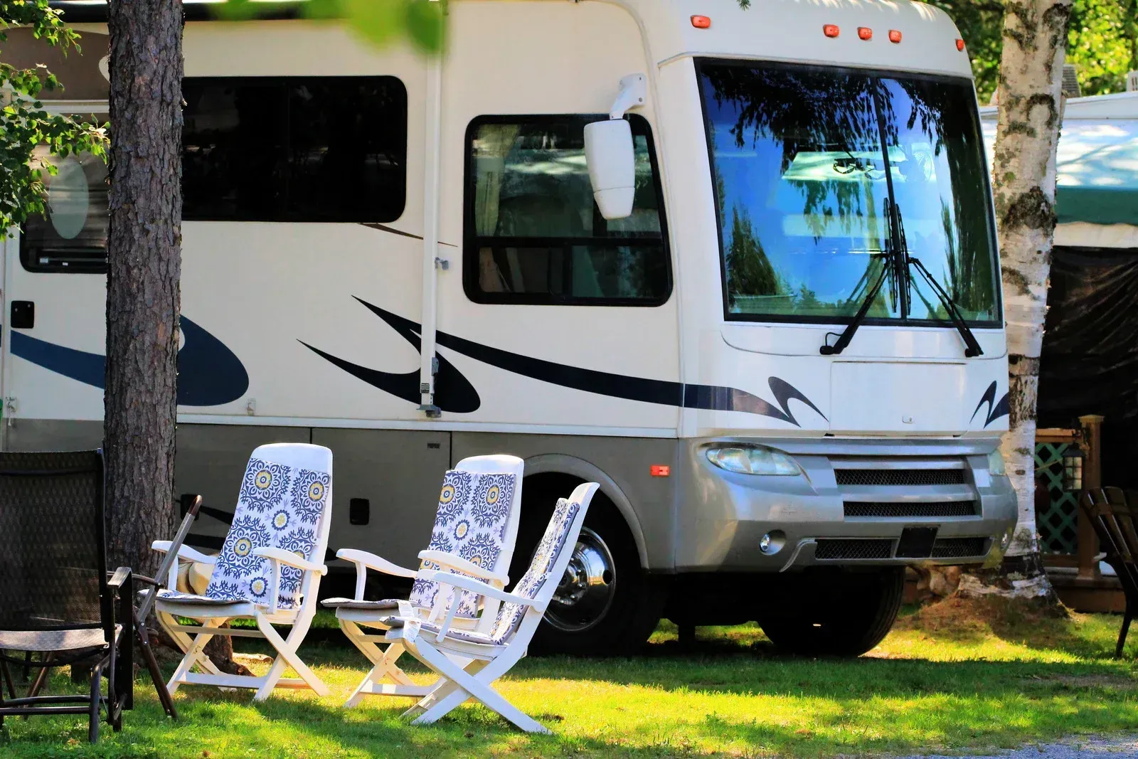 White RV parked on grass beside two patterned folding chairs in a sunny campsite.