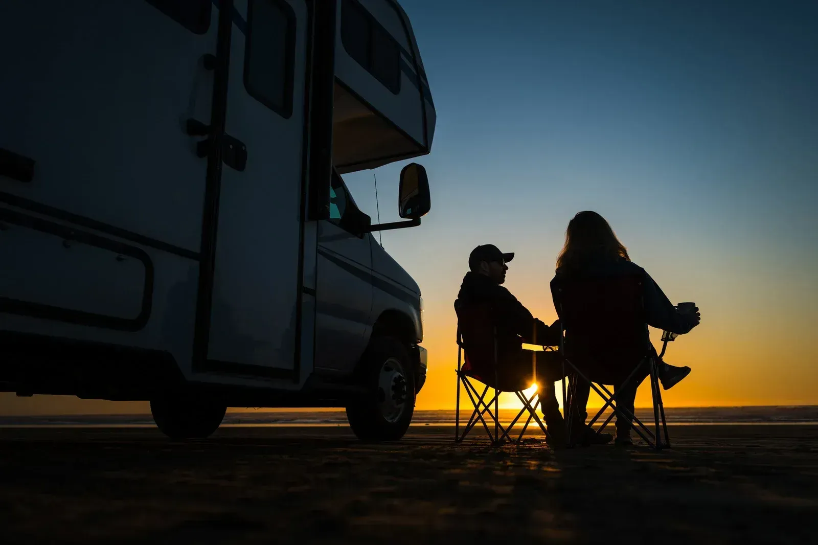 Silhouetted campers seated beside an RV at sunset on open ground