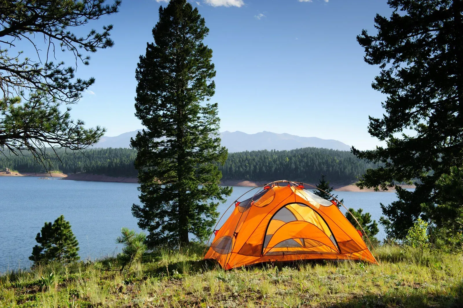 Orange tent by a lake with pine trees and mountains in the background