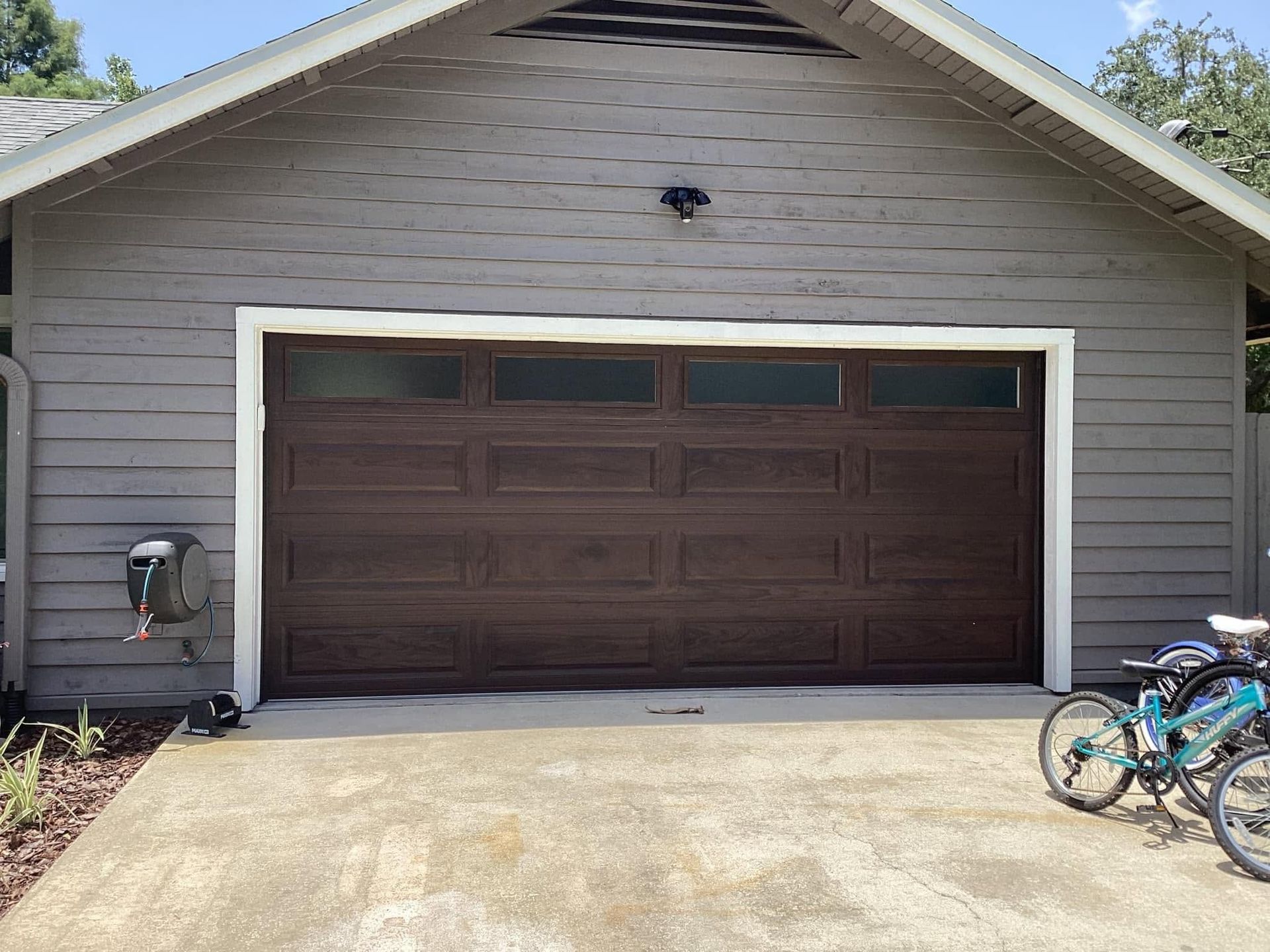 Garage with closed dark brown door, gray siding, and two bicycles parked at the right side of the driveway