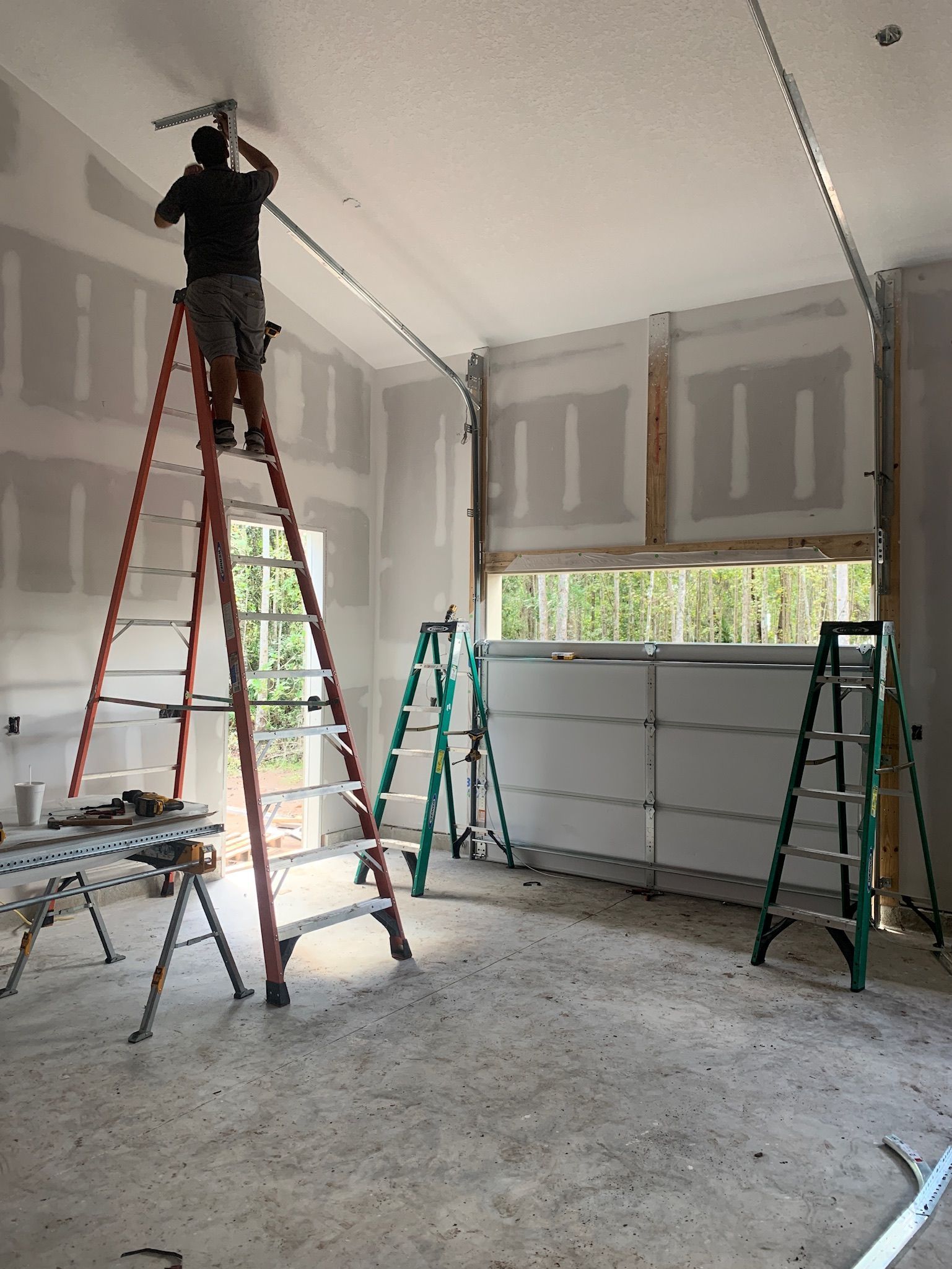 Worker on tall ladder painting drywall in an unfinished room with exposed studs and tools.