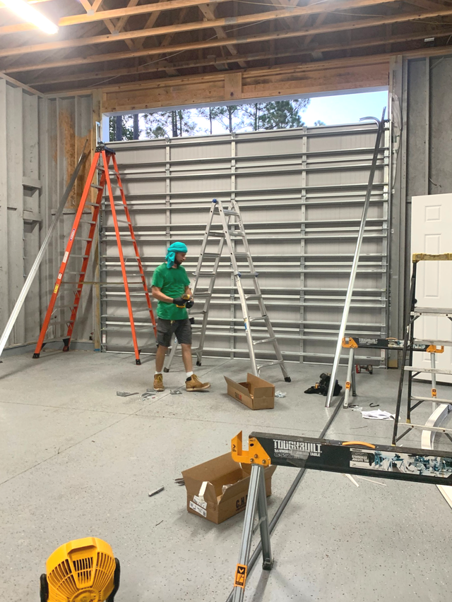 Worker in green hard hat inside warehouse, standing by ladders and stacked wall panels.