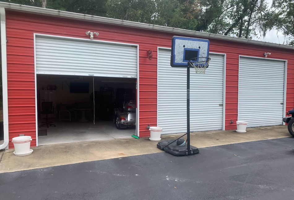 Red storage building with three garage doors, one open, and a portable basketball hoop on the driveway.