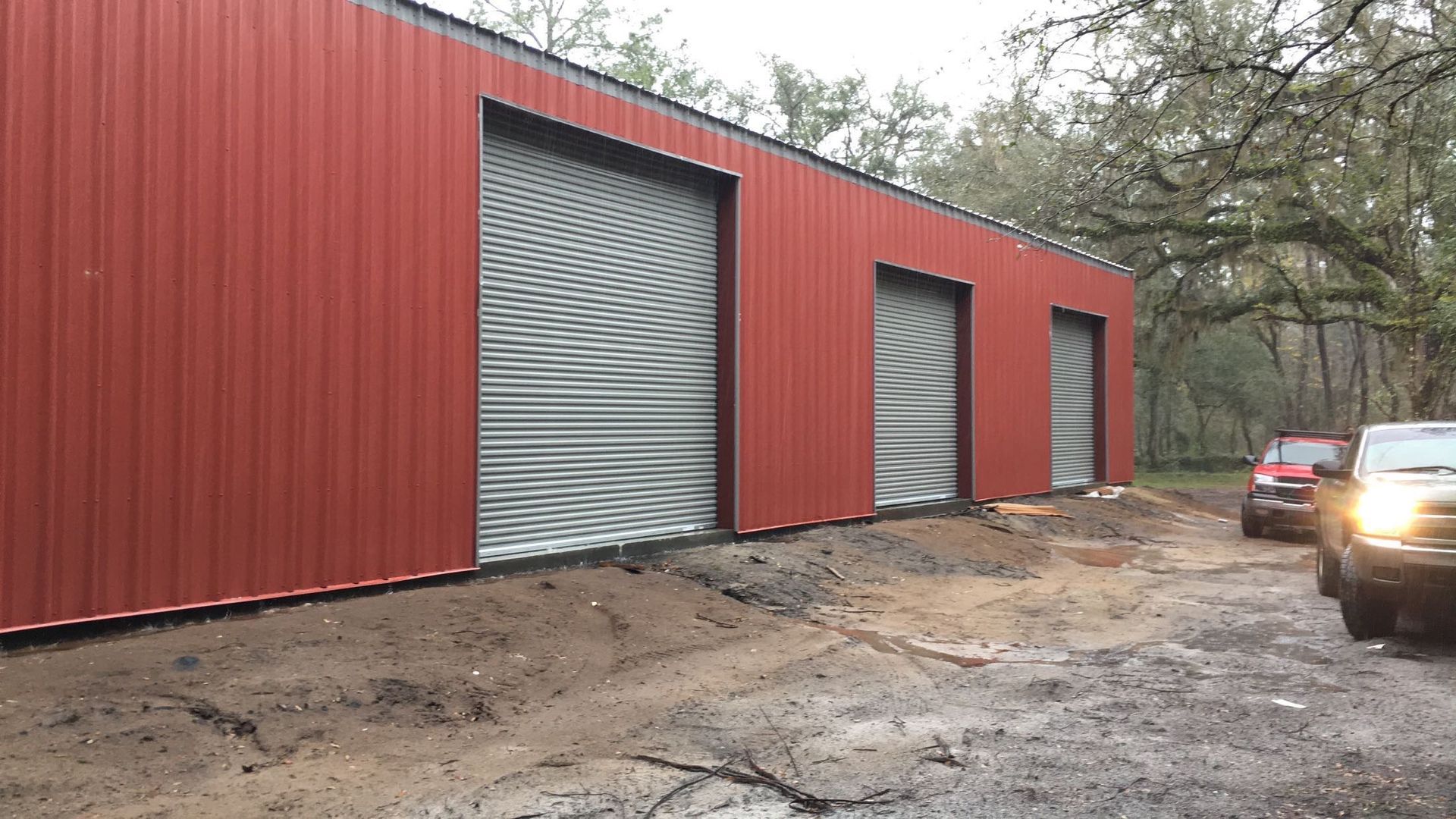 Red metal building with large roll-up doors along a muddy roadside, with a car at right.