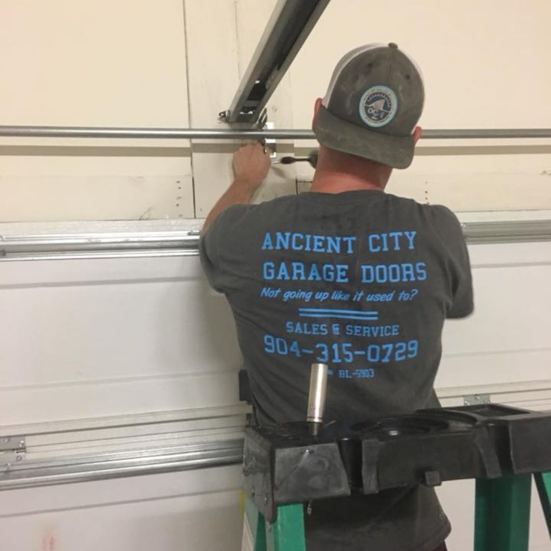 Technician adjusting a white garage door inside a garage, wearing a gray T-shirt with blue company text.