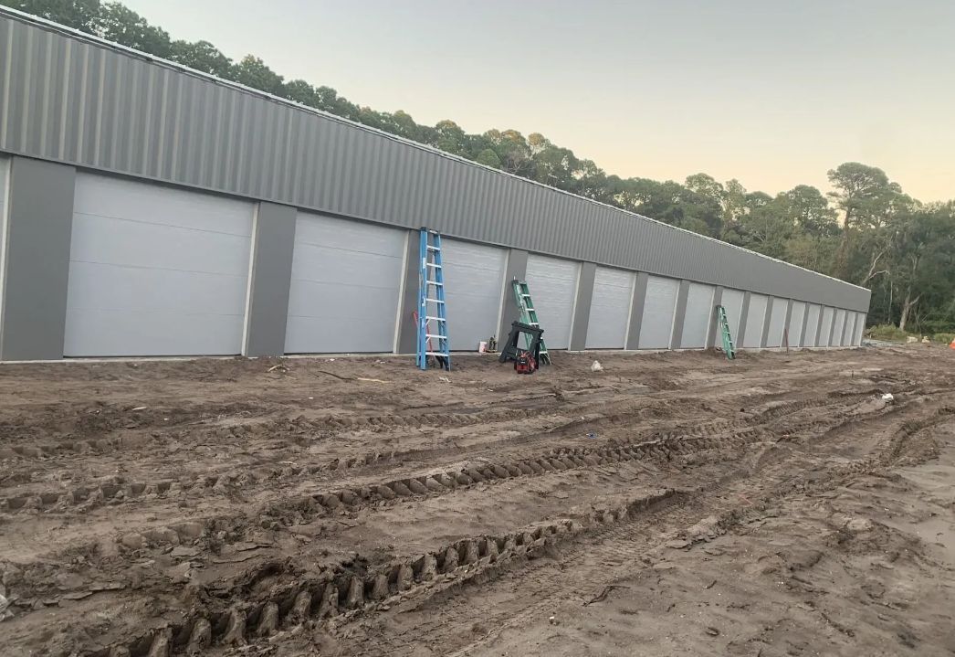 Long warehouse under construction with gray doors, ladders, and muddy ground