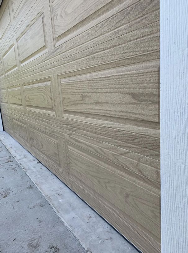 Light woodgrain garage door beside a concrete driveway and pale blue wall