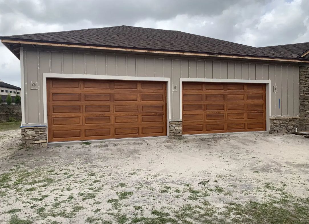 Two brown garage doors on a white building with a gray roof, set on a gravel lot under cloudy skies