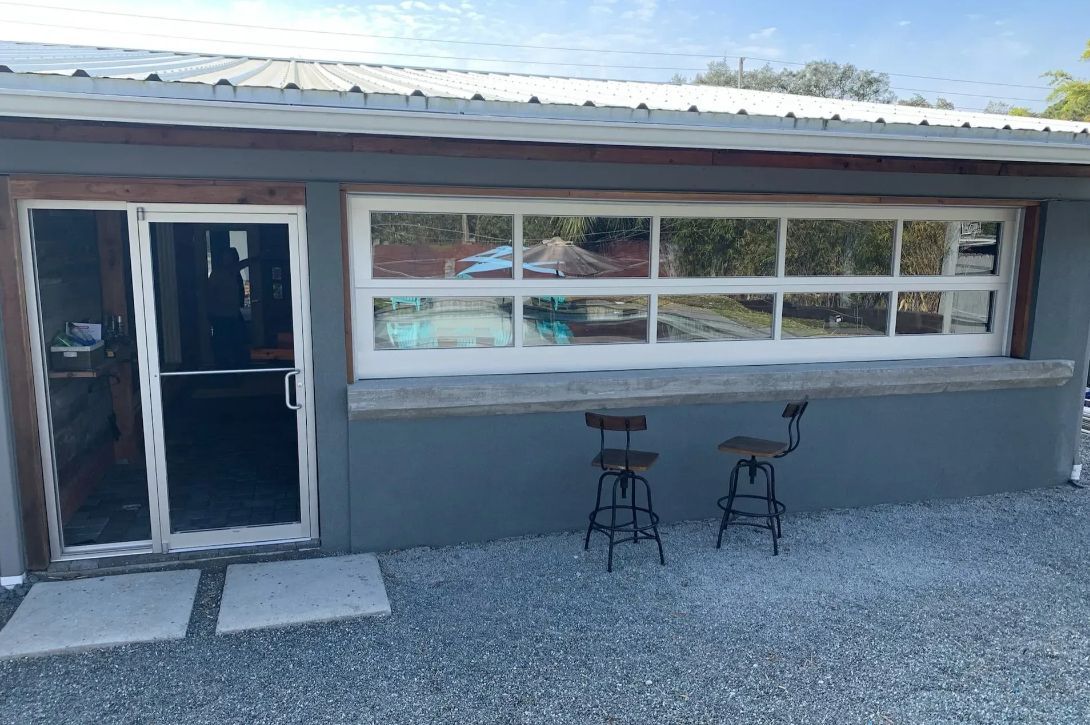 Exterior of a small gray building with a glass door, large window, and two stools on a gravel patio