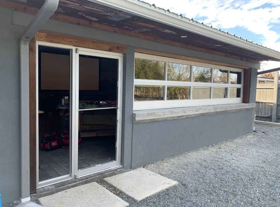 Exterior of a small building with sliding glass doors, long window, and gravel walkway under a sloped roof