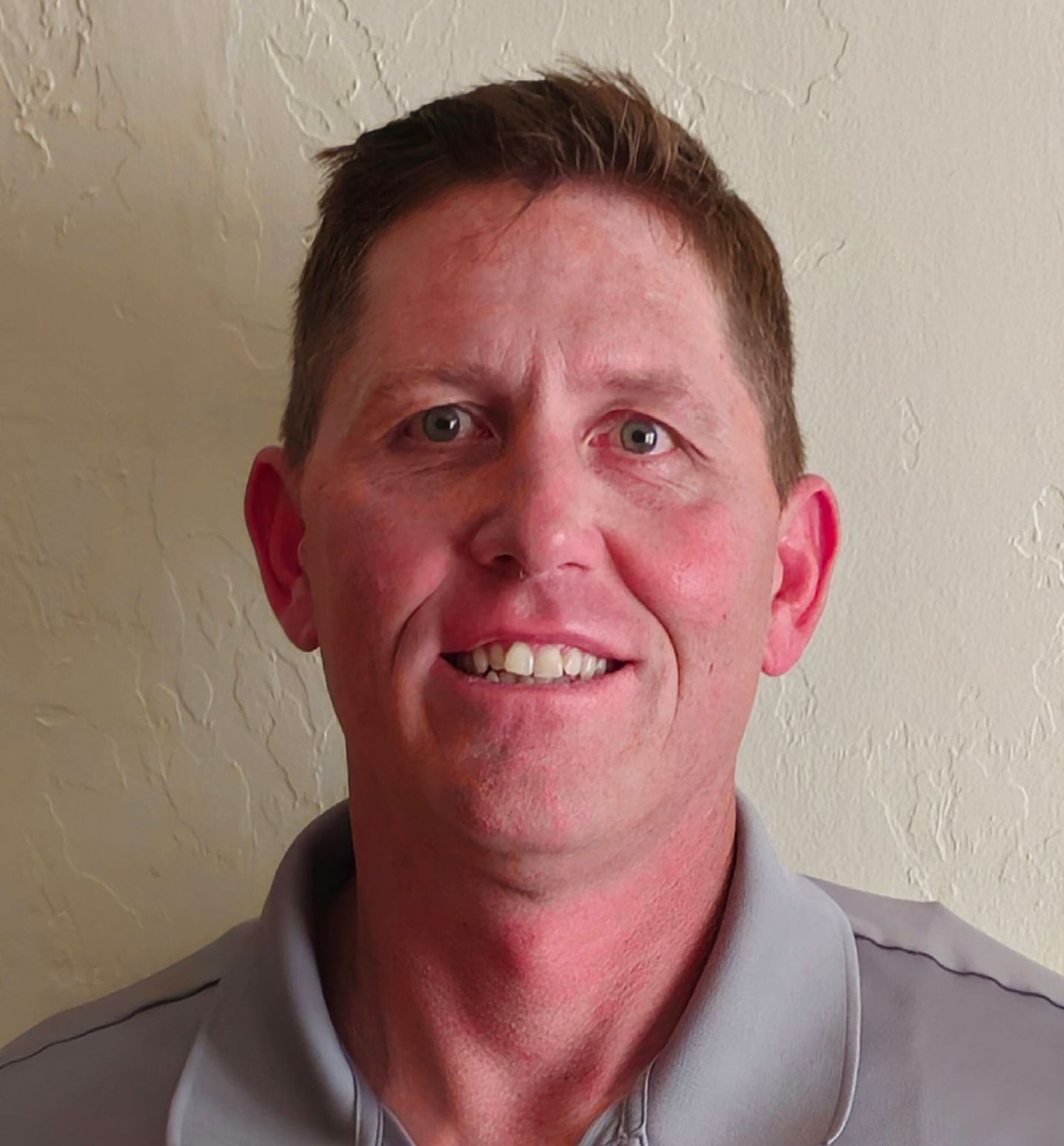 A man in a grey shirt is smiling in front of a white wall