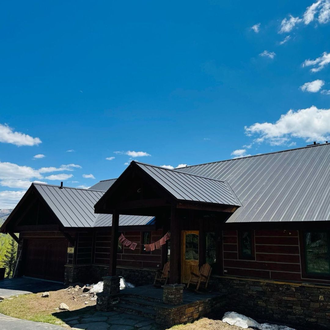 A log cabin with a dark metal roof, stone foundation, and covered entryway against a bright blue sky with scattered clouds.