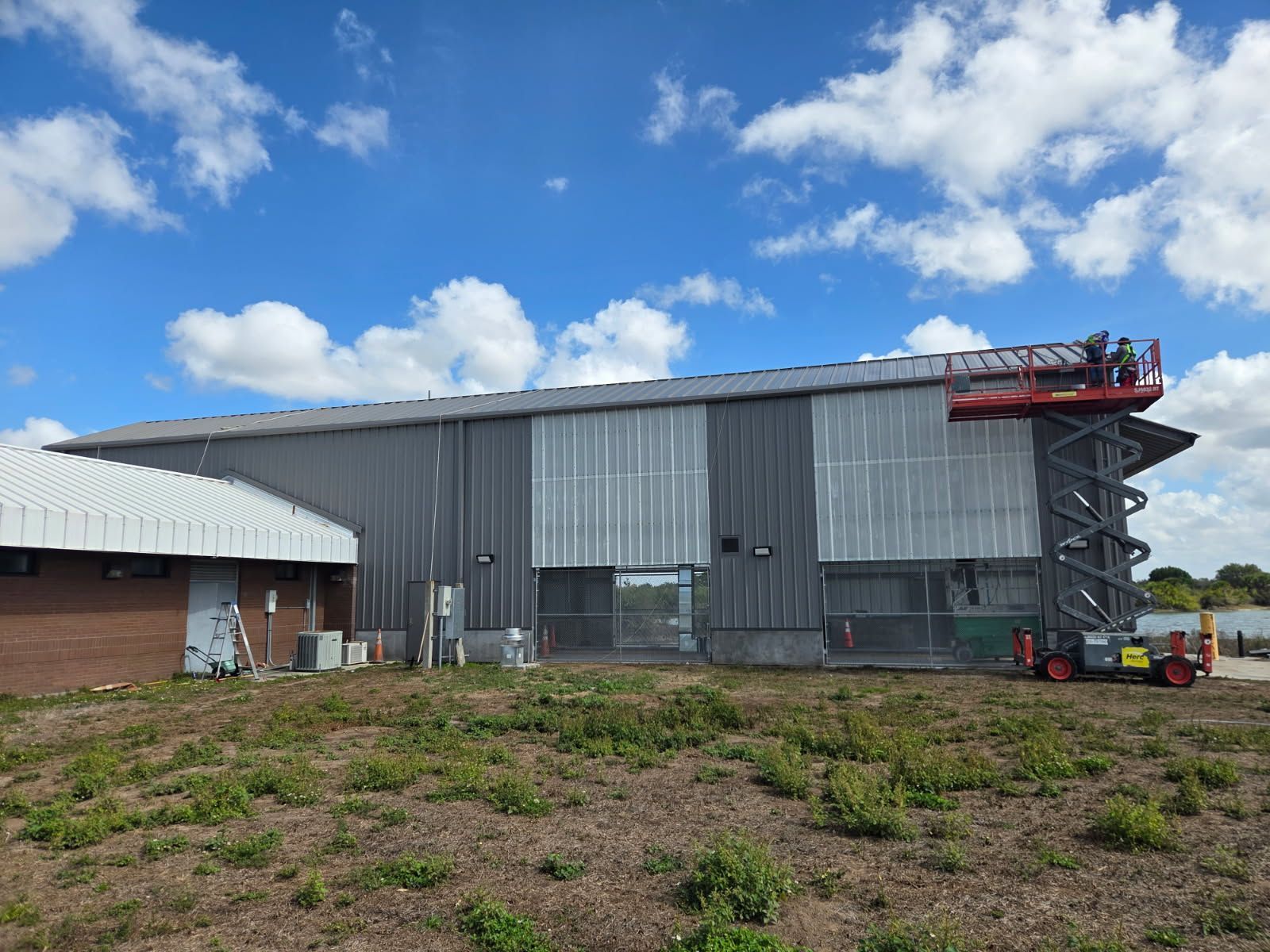 A metal building with a lift platform; two people work on the roof under a blue sky.