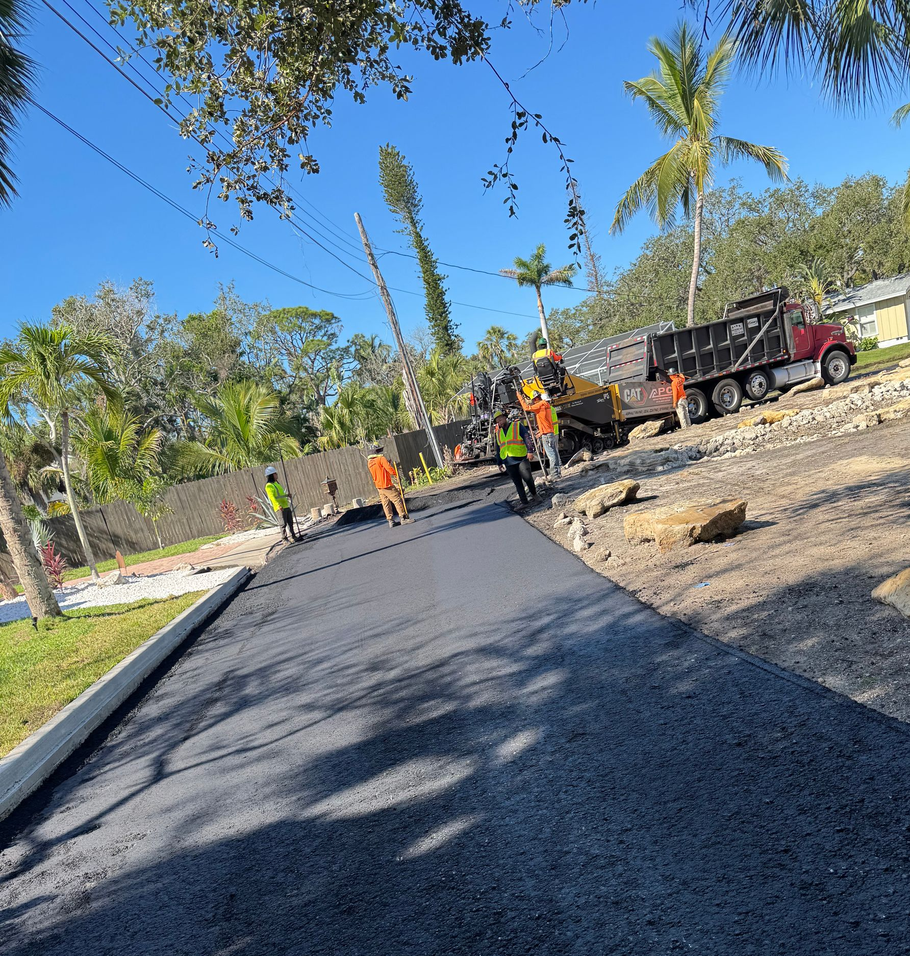 Road paving in progress; workers in safety vests lay asphalt, dump truck in the background, sunny day.