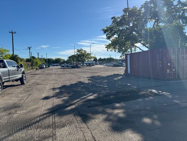 Gravel parking lot on a sunny day with a truck, trees, and some parked vehicles. A red metal container is on the right.