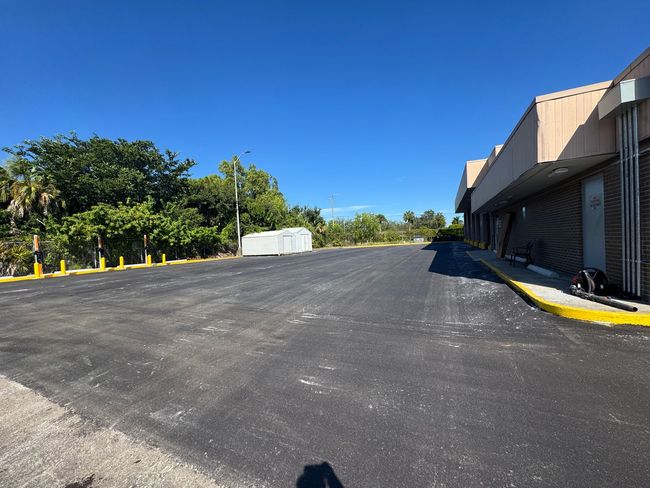 Asphalt driveway next to a brick building and greenery under a clear, blue sky.