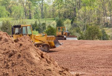 Yellow bulldozers on a construction site, moving red soil. Trees are in the background.
