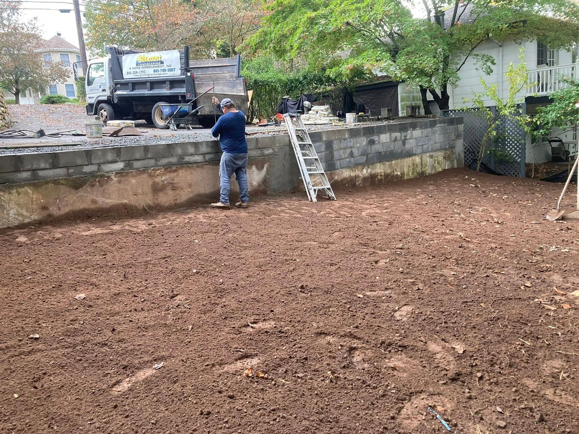 Man standing in a dirt yard next to a concrete retaining wall, with a dump truck and ladder visible.