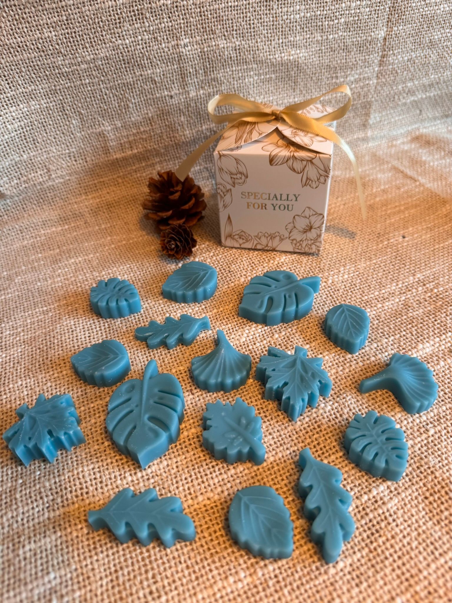 Blauwe bladvormige knutselwerkjes op jute naast een kleine dennenappel en een geschenkdoos met lint.