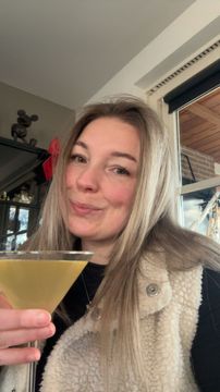 Vrouw in een beige jas, glimlachend en met een martiniglas in haar hand, staat binnen bij een raam.