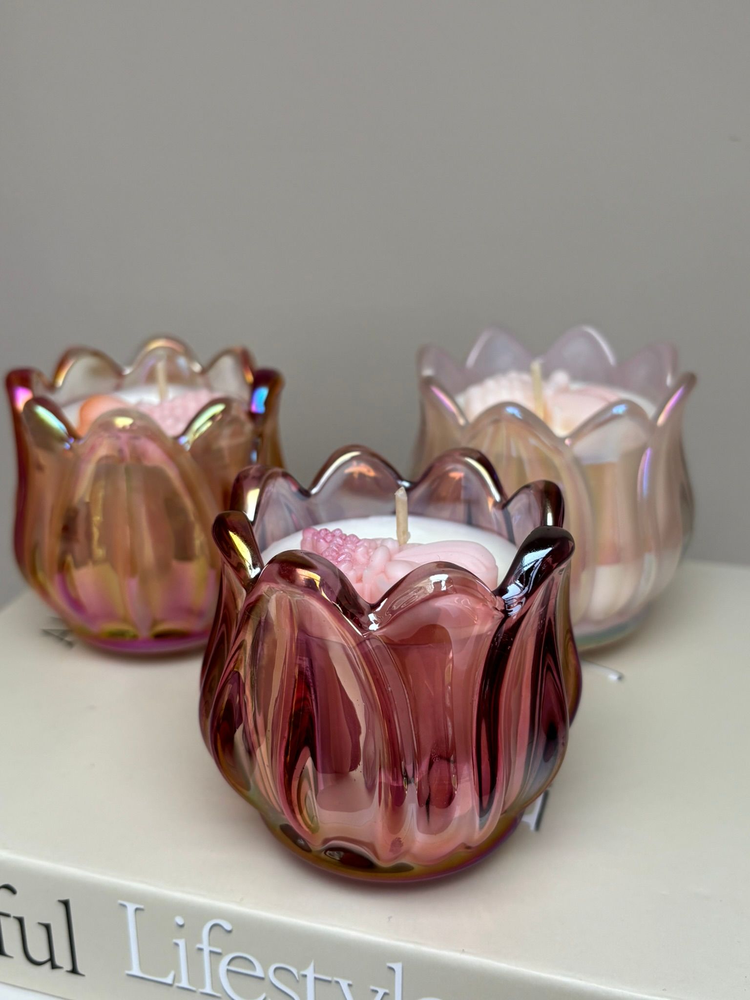 Drie iriserende, lotusvormige glazen kaarsenhouders in roze, amber en wit op een plank.