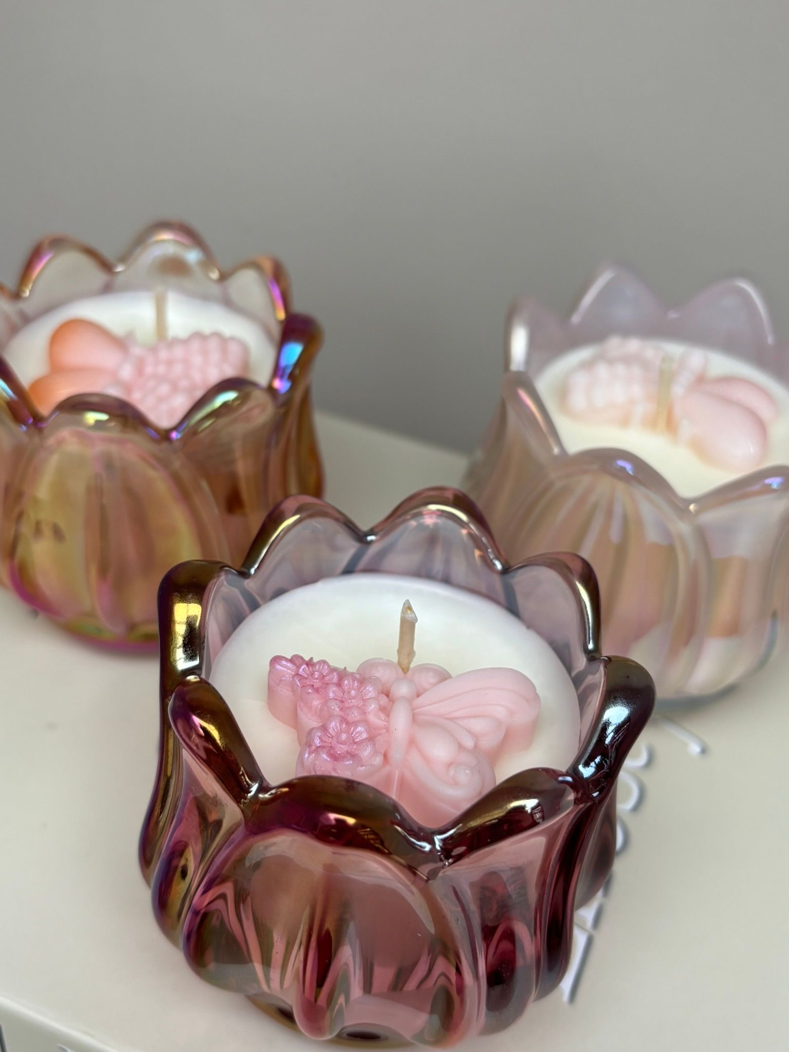Drie iriserende, lotusvormige glazen kaarsenhouders in roze, amber en wit op een plank.