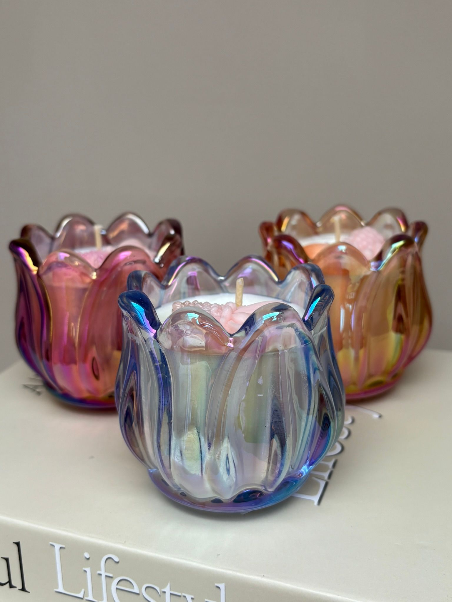 Drie iriserende glazen kaarsenhouders in roze, blauw en goud op een lichte ondergrond.