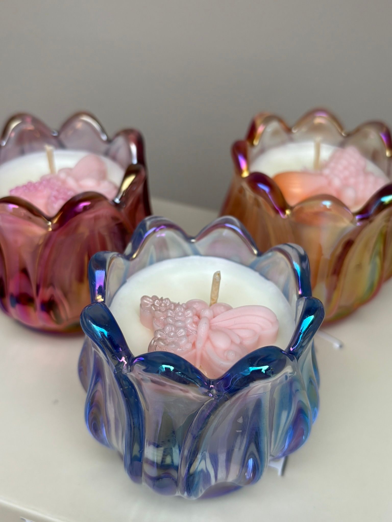 Drie iriserende glazen kaarsenhouders in roze, blauw en goud op een lichte ondergrond.